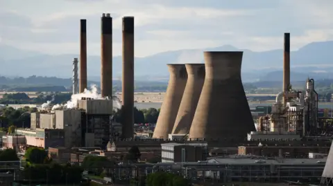 An oil refinery with three large tall chimneys to the left of the photo and three large wide chimneys in the centre