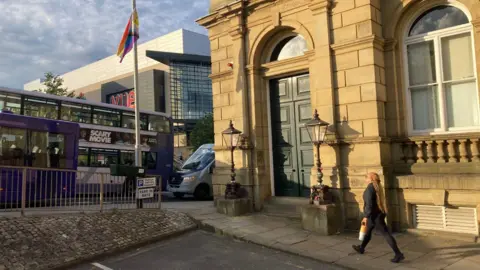A person with small flags in her hairband looking up at a lamppost with a multi-coloured flag tied to it (taken 22 June)