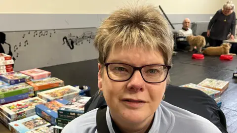 John Devine/BBC Lucinda Stannard has sandy coloured spikey hair with dark framed glasses on, she has a grey fleece top on and is on a mobility scooter, behind her is astage with lots of jigsaw puzzle boxes on.