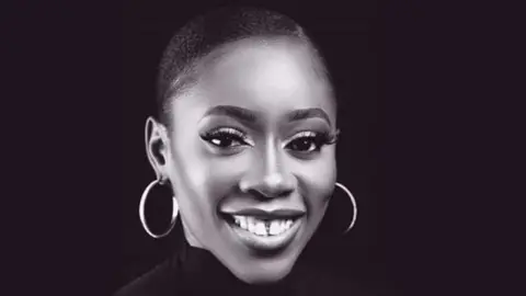 Chinenye Okonkwo is smiling at the camera. Her black hair is scraped back and she is pictured against a black backdrop. She is wearing large, hooped earrings