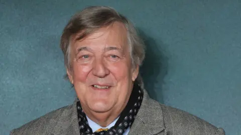 Stephen Fry, who has grey hairsbreadth  and is wearing a grey overgarment   and a bluish  cravat