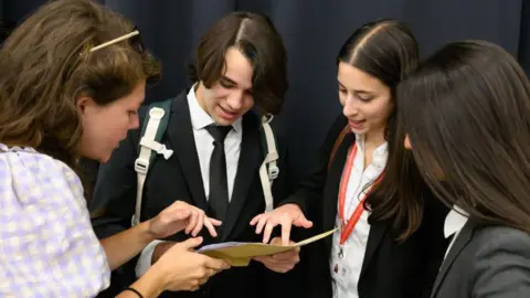 Getty Images Three school pupils wearing uniforms consisting of black blazers and white shirts are looking down at one pupils page showing their A-Level results. 