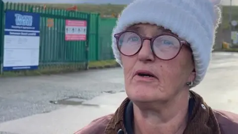 Alison Roe is wearing a white beanie hat and plum-coloured large spectacles to match her jacket.