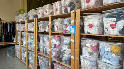 A row of shelves stacked with clear plastic boxes filled with clothes. The boxes are labelled with children's age ranges, as well as the price of the garments.