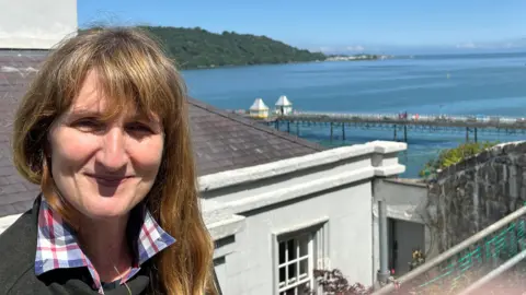 Alwen Gwyn looking at the camera with the sea and Bangor pier behind her