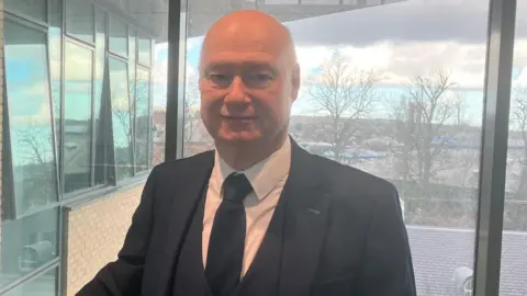 Paul Bartolomew stands in front of a window on the second floor of the university in Derry. A city scape can be seen behind him. He is wearing a dark suit, white shirt and dark tie.