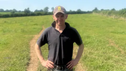 Rob wears a dark green polo shirt and a baseball cap and has his hands on his hips. He is standing in a grassy field with hedgerows and trees in the distance. 