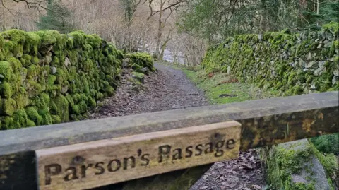 Alan Cleaver A wooden gate that has a sign on it that reads 'Parson's Passage'. Behind it is a stone path, flanked on both sides by a stone, moss covered wall.