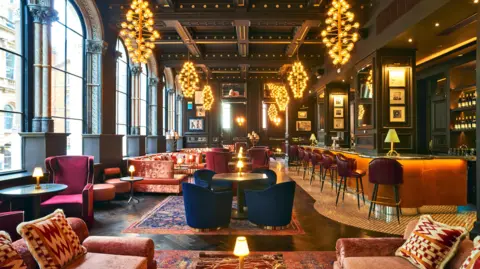 Grand Hotel Image shows a darkened room with eight chandeliers suspended from the ceiling, there are coral coloured sofas and blue velvet chairs arranged around tables, with dark maroon bar chairs up against an orange bar