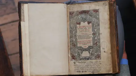 A man in a grey suit with grey hair holds an antique-looking book. The photo is captured from above so the viewer looks down on the scene. The man is stood on a wooden floor with a red carpet laid down. The book has a beautiful Celtic design on the right hand page and calligraphy. The paper in the book is yellowed and spotted with age.