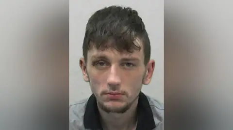 Mugshot of Hetherington. He looks scruffy with a long brown fringe and a wispy goatee beard.