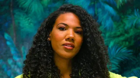 Eden Mullane Eden Mullane smiles at the camera. She has long, black, curly hair. She is standing against a wall that has been painted with green forest leaves.