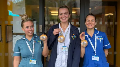 Three women in nurse's uniforms stand in front of a set of glass doors. They are smiling and holding up cardboard baubles with the words 'Light up a Life 2025' on them. 