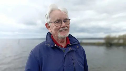 Paddy smiling at the camera. He has grey hair and is wearing glasses and a red polo shirt with a navy jacket zipped up over it.