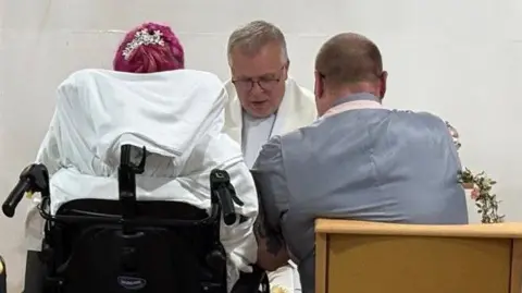 NUH An image of Carla and Craig during their wedding ceremony. The image, taken from behind the couple, shows Carla in a wheelchair with a neck and head support, and Craig sitting on her right-hand side in a chair. Father Gary can seen facing them.