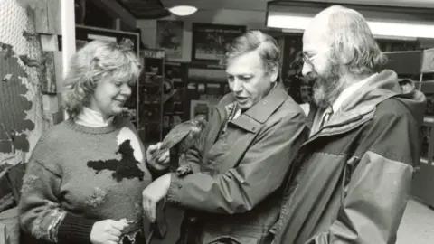 Sir David Attenborough pictured at the Surrey Wildlife Trust centre at Nower Wood near Leatherhead. The picture, taken in 1985, is in black and white. Sir David is stood beside two people at the centre, and has a bird on his left arm.