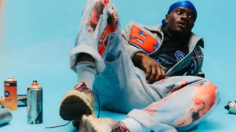 V.I.C wears a blue durag, grey jacket and baggy jeans with turn ups. He's leaning back on his elbow in a light blue studio surrounded by spray cans.