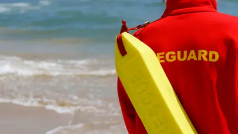 Close up of the back of a lifeguard wearing a red top with yellow letting that says "lifeguard". In the background you can see the sea. The lifeguard is wearing a yellow buoy. 