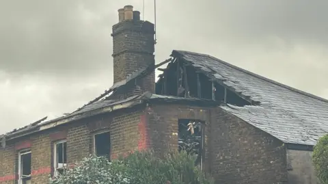 Stephen Huntley/BBC The gutted rooftop of the house, which has lost tiles and had its wooden beams exposed. An intact chimney protrudes through the damage. 