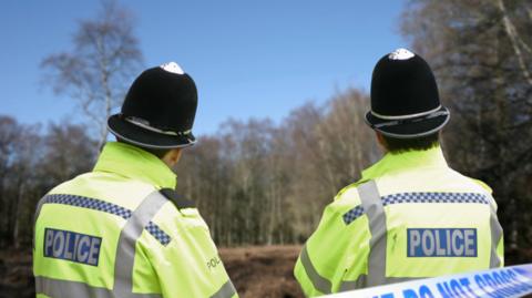 Two police officers with hi-vis police coats on with their backs to the camera looking towards a wooded area 