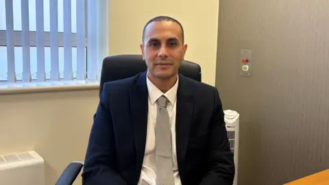 Danielle Malgwi The image shows a man sat in an office wearing a navy blue suit jacket and a grey tie. 