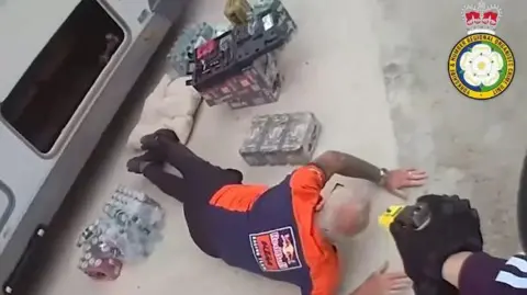 West Yorkshire Police A man in an orange and blue shirt lies on the floor surrounded by cans while the gloved hand of a police officer points an electric stun gun at him.