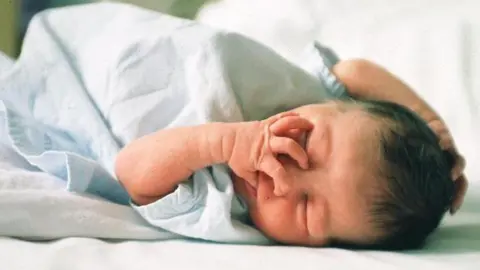 BBC Image of a newborn baby asleep in a hospital maternity ward. The baby has dark hair on its head.