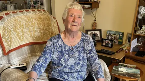 Margaret Quinton sat in a comfy armchair in her living room. She has short white hair and is wearing a blue-and-white paisley patterned top and black trousers. Next to her is a sideboard which has family photos, remote controls and a tea tray on.