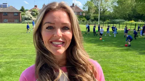 Sam Read/BBC Charley Oldham, who has long mid-brown hair, is wearing a pink top and smiling at the camera. Behind her is a field and children in blue uniforms can be seen kicking balls around.