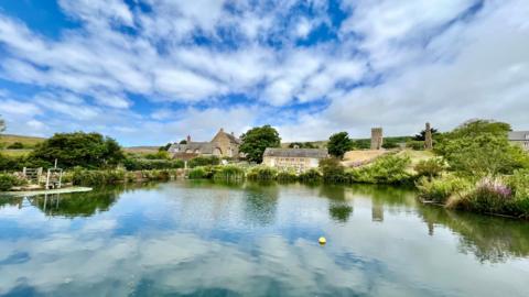 WEDNESDAY -  A body of blue water in the foreground with white clouds reflected. On the far bank are four traditional stone houses and a church tower. The blue sky is streaked with fluffy white clouds on a sunny day. The banks of the water are green with plants and trees.