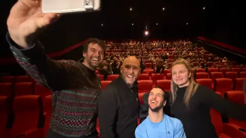 Jimmy Simpson, who is wearing a bright blue top and sitting in wheelchair, poses for a selfie picture which is being taken by his surf instructor, Nick, who has a white and silver phone and is wearing a striped patterned jumper. Cinemagoers can be seen in the seats behind them.