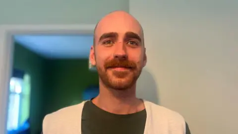 Mid-shot of Tobias Stewart. He has short hair, a beard and a nose piercing. He is wearing a white cardigan over a dark green t-shirt.