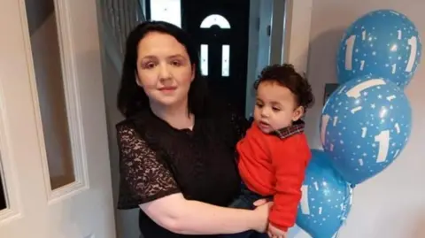 Mandy Kelly A woman and a child. The child is wearing a red jumper. The woman is wearing a dark top and has dark hair. Next to the chid, there are three blue balloons with the number '1' on them.