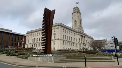 A white stone built building has a large clock tower and dozens of large windows. Outside is a public square, steps and a large piece of public art which looks like a rusty comb.