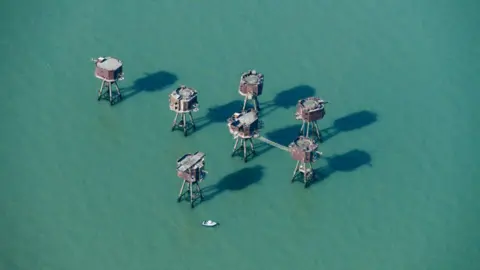 Getty Images An aerial view showing the Redsands forts in the middle of the sea. There are seven of them and they are octagonal shaped structures rising out of the water on long metal legs. They are brown and rusting and a small boat is in the water near to them.