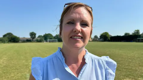 BBC/Gavin Kermack A woman with reddish-blonde hair and sunglasses perched on top of her head, wearing a light blue blouse and standing on a football pitch with a goal in the distance.