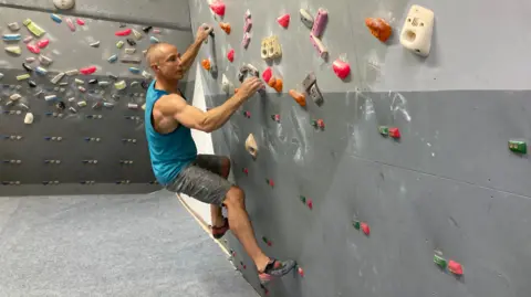 Ben Mitchell Neil Gresham climbs on a dark grey and light grey climbing wall, on which there are lots of grips or holds in bright colours. He has a shaved head and is wearing a blue vest and grey shorts with climbing shoes. He appears not to be far from the ground but is holding himself up by such small holds that the tension in the muscles on his upper arms and forearms is very defined.