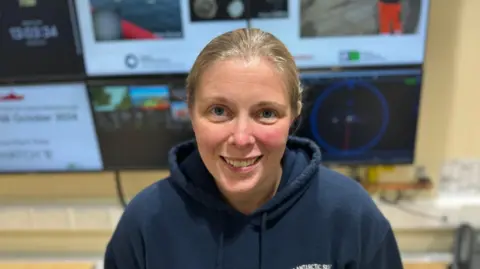 Elliot Deady/BBC A smiling Natalie Ensor looking straight at the camera. She is wearing a blue hoodie and is standing in front of several computer monitors.