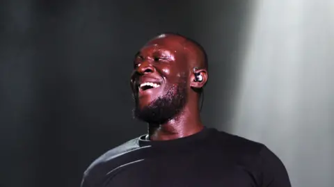 Sarah Jeynes/BBC Stormzy performing at Radio 1's Big Weekend. He is smiling on stage while sweating. He is wearing a black jumper. He has an earpiece in, short buzz-cut hair and a dark beard. Behind him is a spotlight and plain black and grey background.