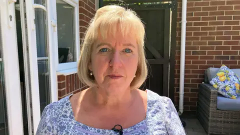 Catherine O'Sullivan pictured in her garden. She has short blonde hair and a fringe and is wearing a blue and white floral top with a microphone pinned to the neckline.