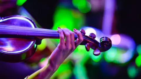 A close up image showing part of a female musicians hand and arm as she holds a fiddle. There are blurry bright lights in the background.
