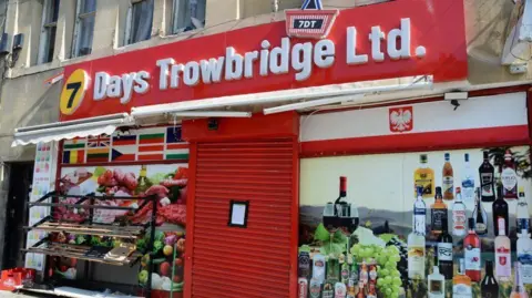 A shop front with a sign over the entrance with the words "7 Days Trowbridge Ltd." written on it. The shop appears closed with the metal shutters down and a notice pinned to the outside.