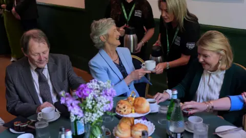 PA Media A woman with short grey hair, wearing a blue suit and navy top turns to her left and laugh's as she receives a cup of tee from a long-blonde haired woman in a dark-green polo top. 