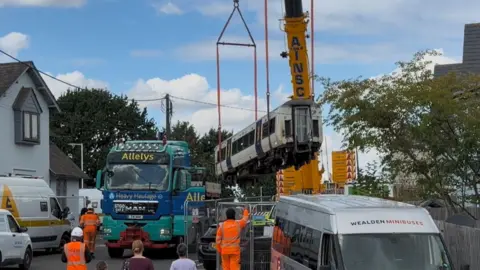 A crane carrying one of the derailed carriages as a small group of members of the public and workers in hi vis jackets and hard hats watch. A few vans and vehicles are parked to the sides and there are trees and housing nearby.