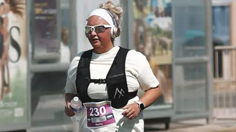 PA Media A picture of Holly running in a race. She is wearing a white vest, a black weighted vest, white headphones and sunglasses and has a purple race number with white 230 pinned to her t-shirt. 
