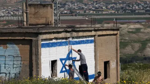 Israeli right-wing activists paint an Israeli flag on the side of an abandoned building in the Jordan Valley, in the occupied West Bank, with neighbouring Jordan visible in the background (11 March 2024