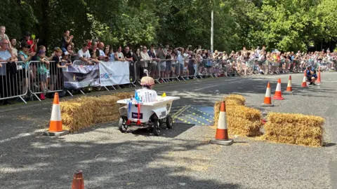 A man sat in a kart made out of a bath tub about to go over a small black ramp in between some straw bales. Crowds of people are gathered behind metal railings to watch. 
