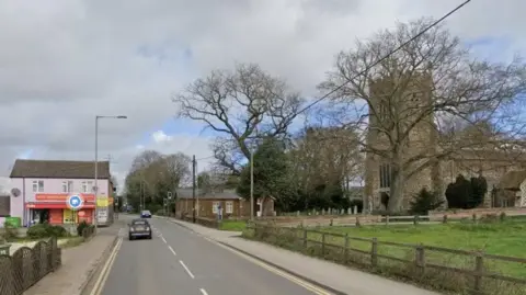 Google maps view of West Winch village. There is a road running through the picture with a pink house to the left and the village church to the right 