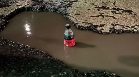 Jason Conyers A full 500ml bottle of Coca Cola sits in a water-filled pothole. The water is up to the bottom of the label, showing the depth of the hole and water. The hole is surrounded by crumbling concrete edges.