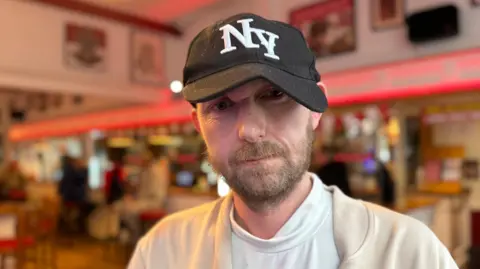 Chris Lewis wears a black New York cap and a brown jacket over a white shirt. He has a light brown beard.